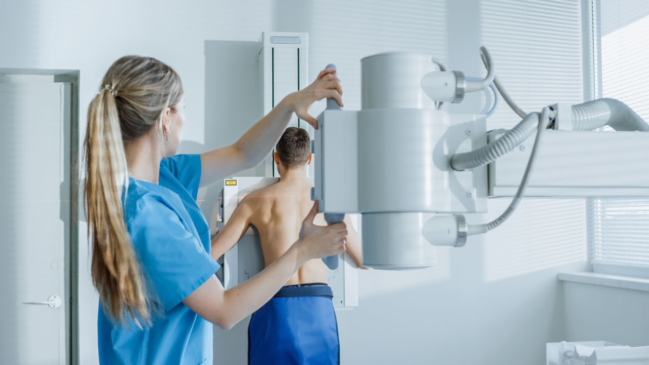 In the Hospital, Man Standing Face Against the Wall While Medical Technician Adjusts X-Ray Machine For Scanning. Scanning for Fractures, Broken Limbs, Chest, Cancer or Tumor. Modern Hospital with Technologically Advanced Medical Equipment.,In the Hospital, Man Standing Face Against the Wall While Medica