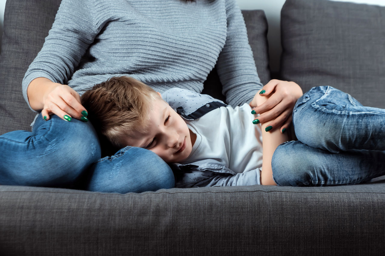 The boy is lying on the couch with an abdominal pain near his mother. The concept of custody, parental care, stomach problems, food poisoning, problems in children.,The boy is lying on the couch with an abdominal pain near his mo
