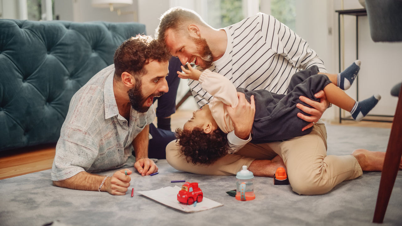Loving LGBTQ Family Playing with Toys with Adorable Baby Boy at Home on Living Room Floor. Cheerful Gay Couple Nurturing a Child. Concept of Diverse Childhood, New Life, Parenthood.,Loving LGBTQ Family Playing with Toys with Adorable Baby Boy at 