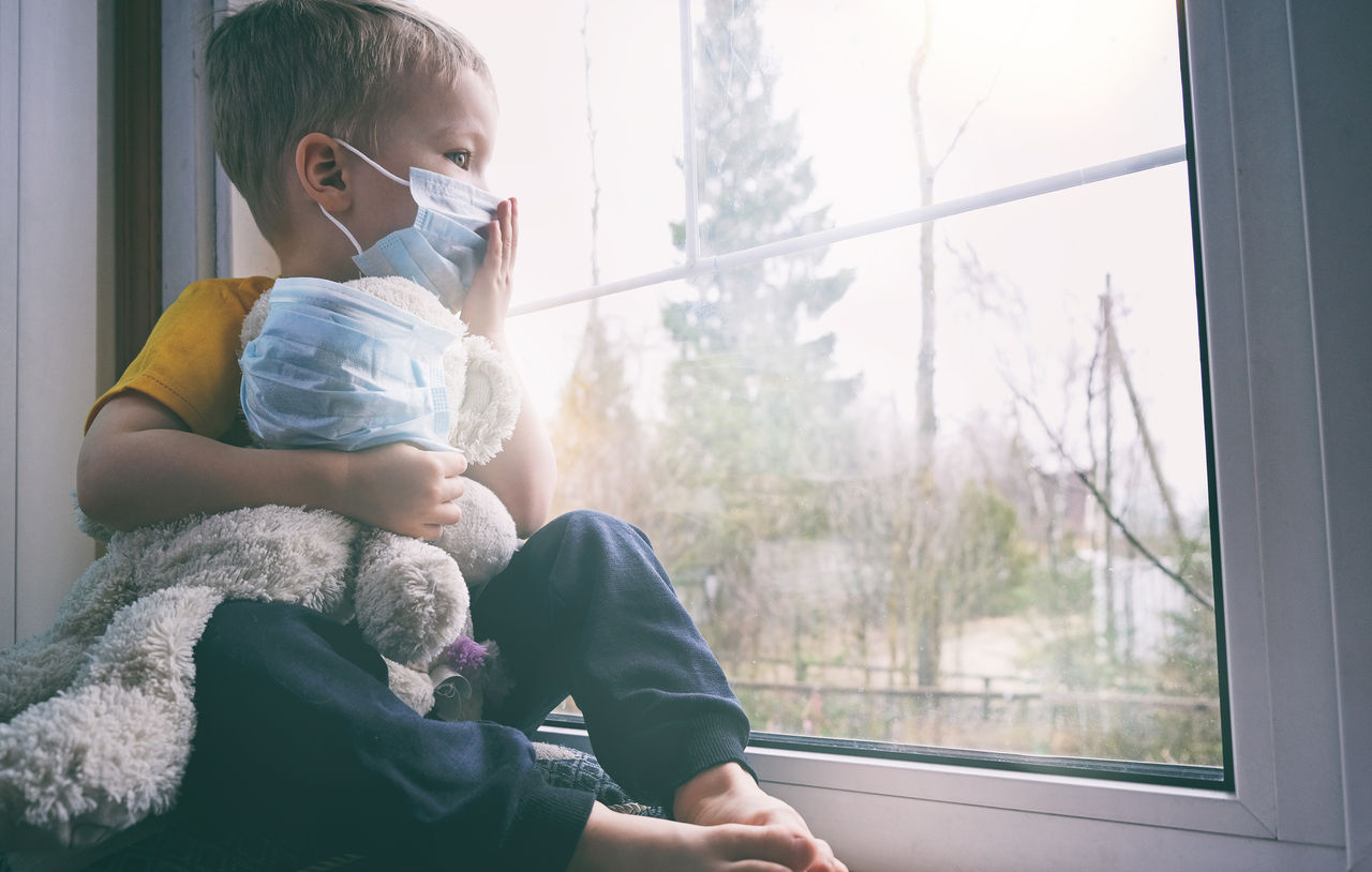 Illness child on home quarantine. Boy and his teddy bear both in protective medical masks sits on windowsill and looks out window. Virus protection, coronavirus pandemic, prevention epidemic.,Illness child on home quarantine. Boy and his teddy bear both in