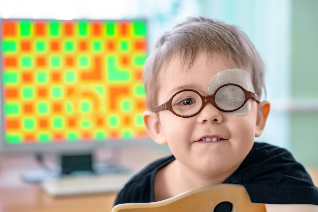 A little boy wearing glasses and an eye patch (plaster, occluder). Apes or grimaces. He undergoes vision treatment to prevent amblyopia and strabismus (squint, lazy eye). Child congenital vision disease problem.,A little boy wearing glasses and an eye patch (plaster, occluder