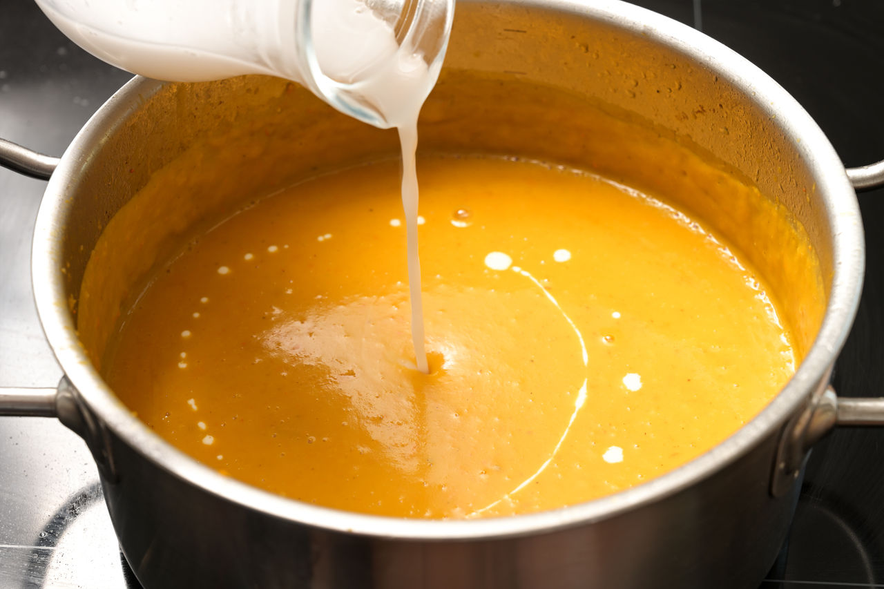 Pouring coconut milk into a vegetable cream soup from squash in a steel pot, vegan cooking in autumn, copy space, selected focus,Pouring coconut milk into a vegetable cream soup from squash in 