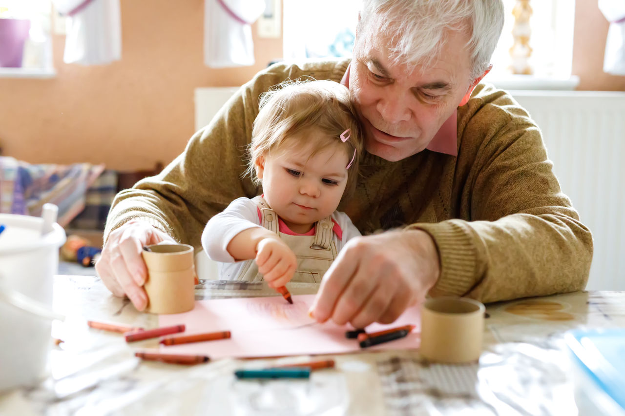 Cute little baby toddler girl and handsome senior grandfather painting with colorful pencils at home. Grandchild and man having fun together,Cute little baby toddler girl and handsome senior grandfather pa
