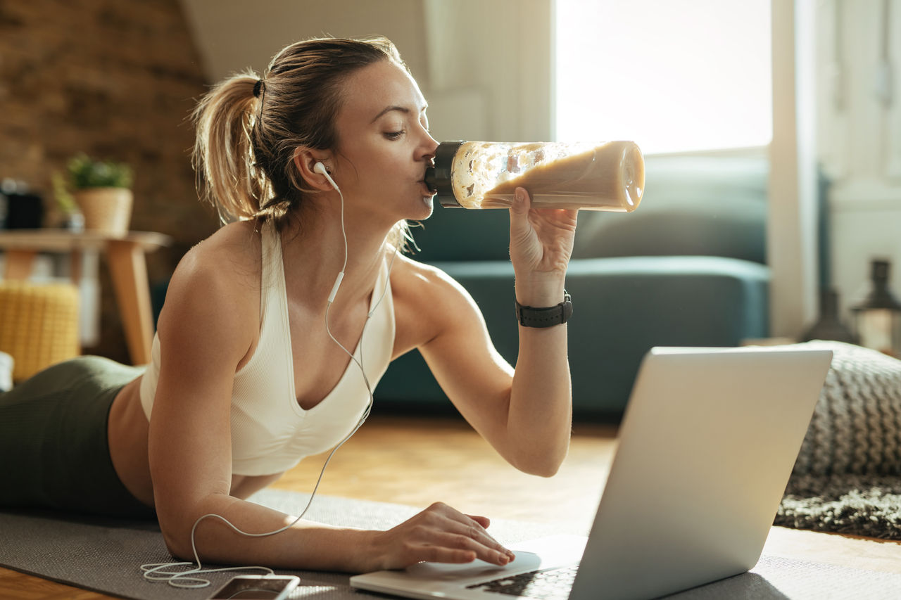 Young athletic woman drinking protein shake while using laptop on the floor. ,Young athletic woman drinking protein shake while using laptop o