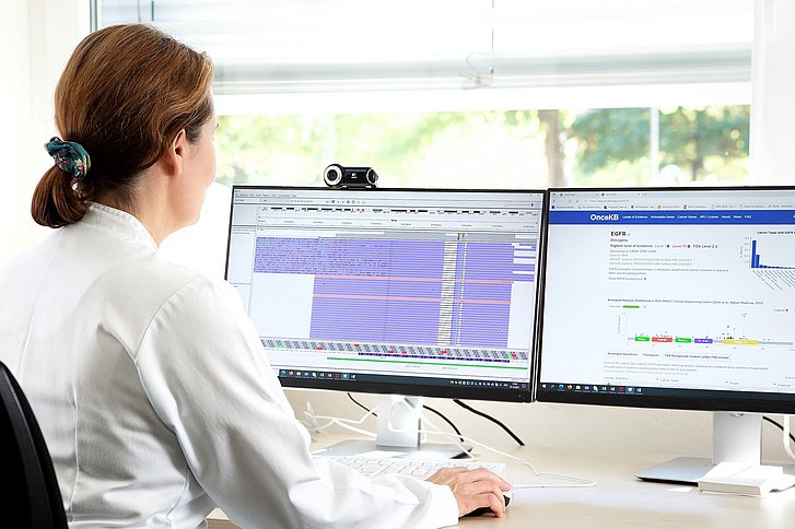 Ärztin in weißem Kittel blickt auf Computerbildschirm, auf dem DNA-Sequenzen dargestellt sind.