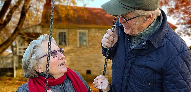 Älterer Mann mit Brille lächelt Frau mit Brille an, die auf Schaukel sitzt