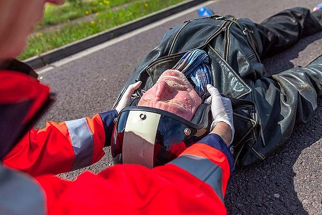 Ein Mann in Motorradkleidung liegt am Boden. Ein Rettungssanitäter zieht ihm vorischtig den Helm ab.