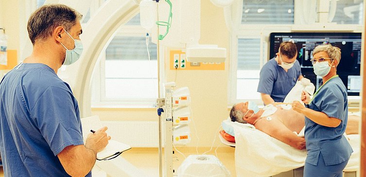 Drei Personen in blauen Kassacks mit Mund-Nasen-Schutz im Herzkatheterlabor