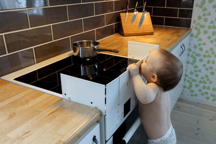Малыш в пеленках пытается дотянуться до плиты.  Вокруг печи установлена ​​защитная решетка.