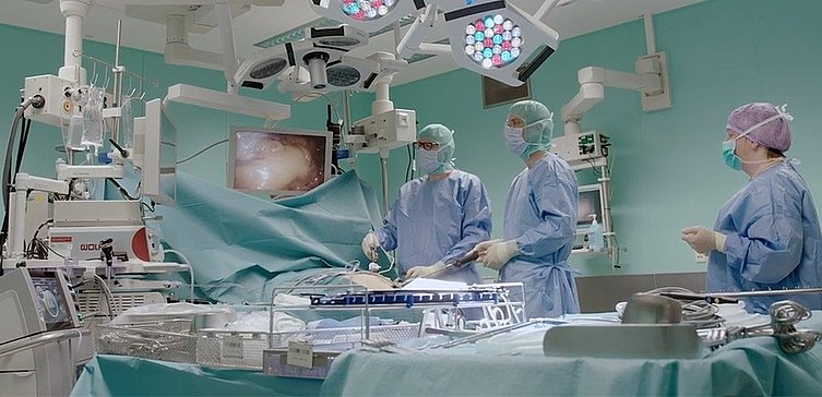 Operationssaal mit drei Operateuren, sowie einem Patient unter OP-Kleidung. Diverse medizinische Geräte.