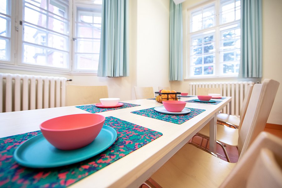 Ein Tisch auf dem sich rosa Schüsseln und hellblaue  Teller befinden. An den Tisch sind drei Stühle gestellt. Im Hintergrund sind zwei große Fenster zu sehen mit blauen Vorhängen.