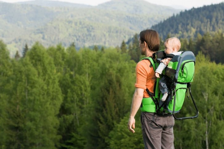 Mann trägt ein Baby in einer Trage auf dem Rücken. Die beiden wandern im Wald. 