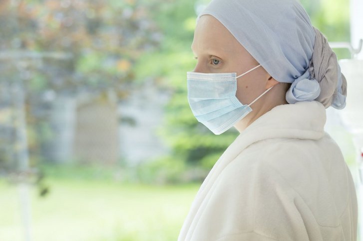 Frau mit Mundschutz und Kopftuch sieht aus Fenter, neben ihr Infusionsständer