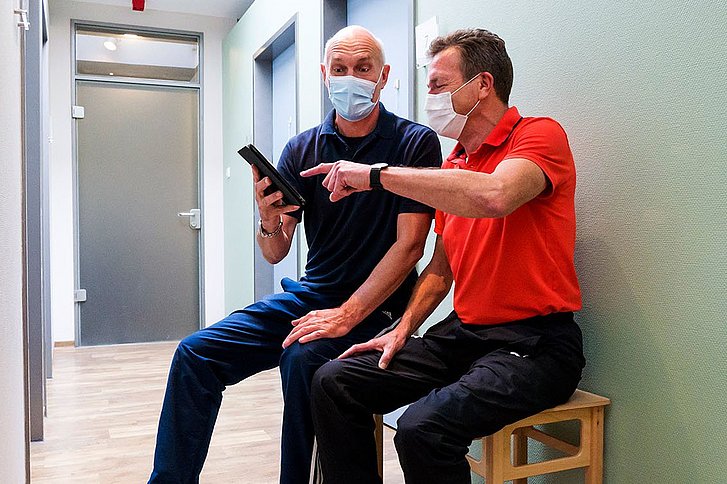 Zwei Männer sitzen im Krankenhaus auf Bank mit Tablet in der Hand