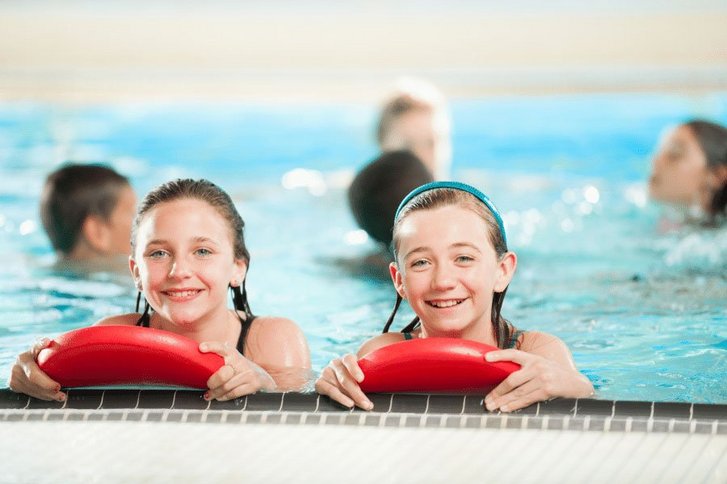 Zwei Mädchen im Schwimmbad. Sie nutzen eine Schwimmhilfe und lächeln in die Kamera. 