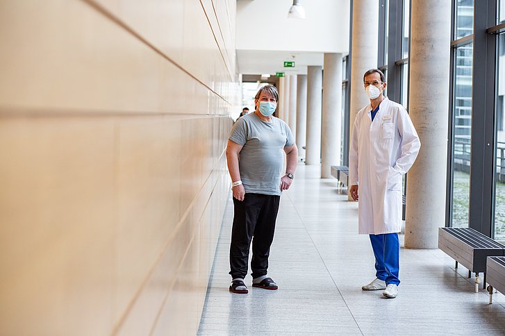 Zwei Männer mit medizinischer Maske stehen auf dem Krankenhausflur, links der Patient, rechts der Arzt.