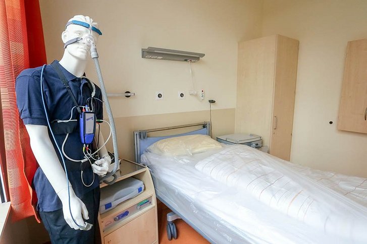 Eine Schaufesnterpuppe steht neben einem Bett. Sie ist verkabelt und trägt einen Patientenkittel. 