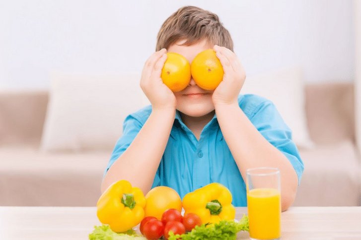 Junge mit blauem T-Shirt sitzt am Tisch. Er hält sich Orangen vor sein Gesicht. 