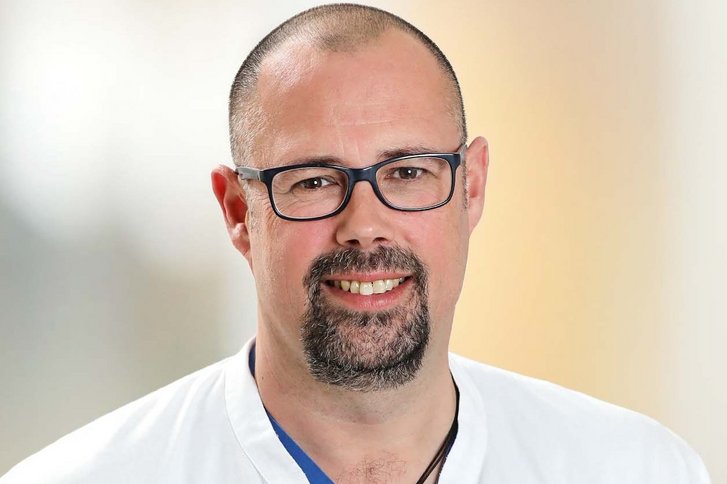lächelnder, dunkelhaariger Mann mit schwarzer Brille und dunklem Bart in blauer Arztkleidung blickt direkt in die Kamera