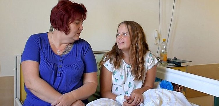 Ein junges Mädchen und dessen Mutter sitzen auf einem Krankenhausbett und sehen sich an. 