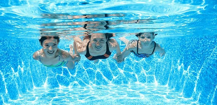 Drei Kinder tauchen im Schwimmbecken und lächeln in die Kamera. 