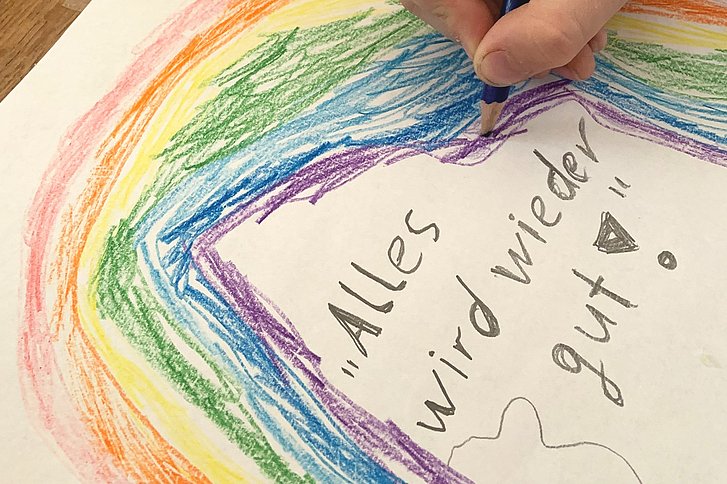 Ein Kind malt einen Regenbogen. Auf dem Blatt Papier steht "Alles wird wieder gut"