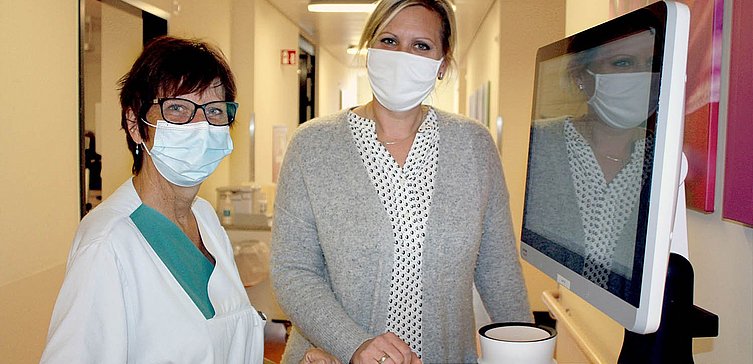 Eine Krankenpflegerin steht neben einer anderen Frau am Visitentisch