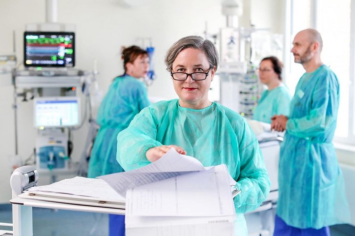 Eine Hygienebeauftragte mit Brille und grünem Schutzkittel steht im Operationsraum und schaut in die Kamera, im Hintergrund behandeln drei Personen einen Patienten