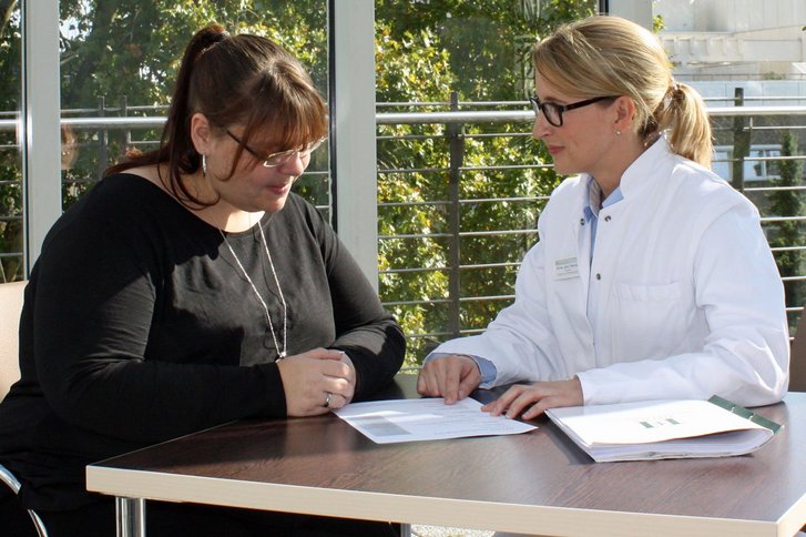 übergewichtige Frau in schwarzer Kleidung neben Frau in Arztkittel am Tisch