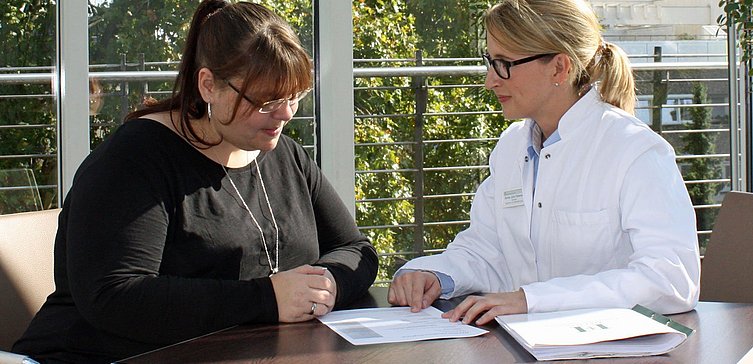 Frau mit übergewicht in schwarzer Kleidung neben Frau in Arztkittel