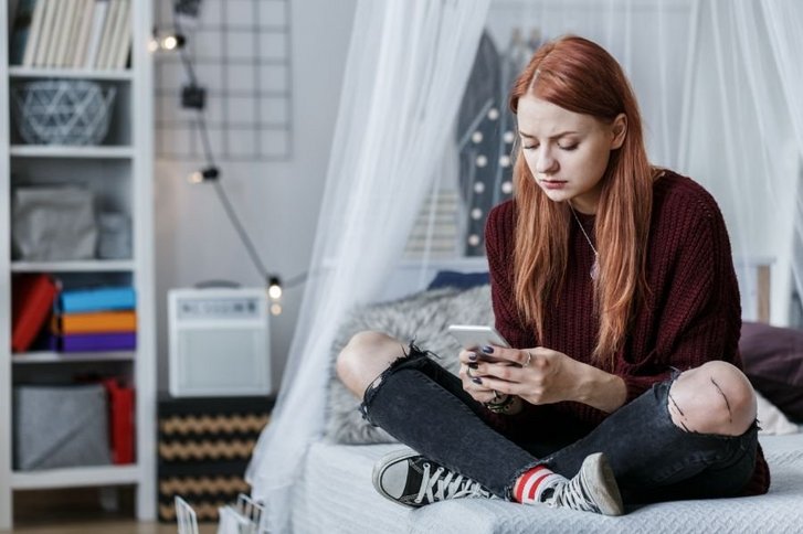 Junges Mädchen mit roten Haaren sitzt auf Bett und blickt auf Smartphone.