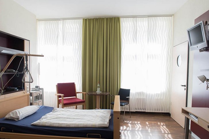 Ein helles Zimmer mit Fenster in dem ein großes Bett und ein Sessel zu sehen sind.