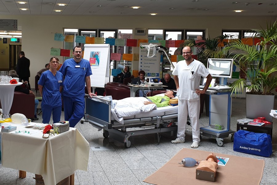 zwei Männer und eine Frau in Krankenhauskleidung blicken in die Kamera und stehen vor Bett mit Reanimationspuppe.