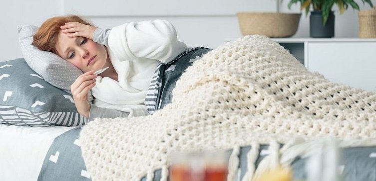 Junge Frau liegt auf Couch unter Decke mit Fieberthermometer in Hand. 
