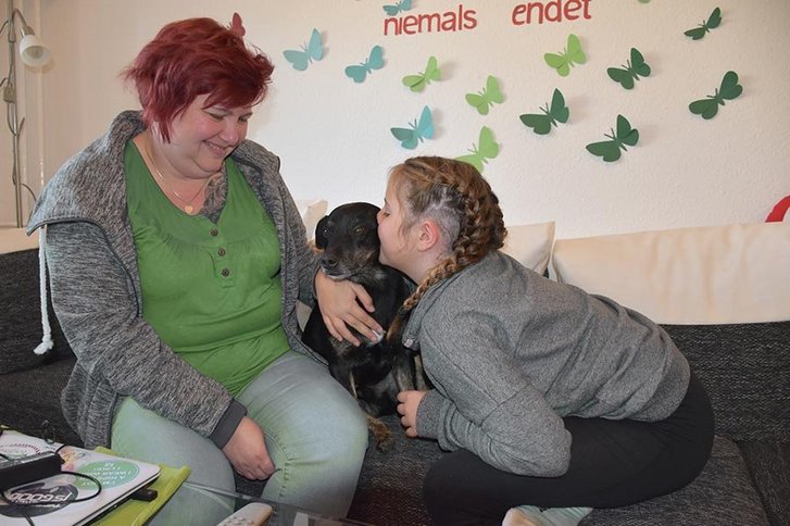 Ein junges Mädchen gibt ihrem Hund ein Kuss auf das Ohr und die Mama lächelt sie an.