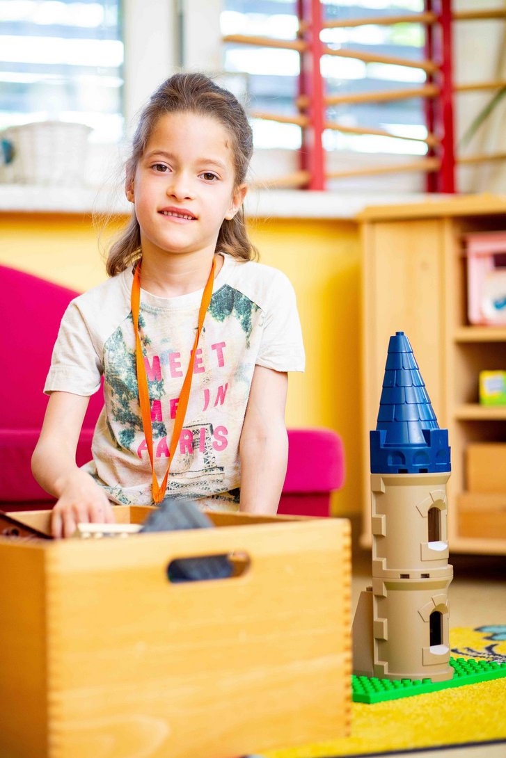 Kleines Mädchen sitz hinter eine Holskiste mit Spielzeug und neben einem Turm aus Bausteinen.