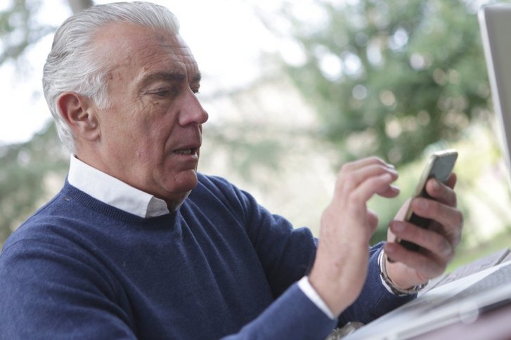 Älterer Mann im Garten am Tisch mit Telefon in der Hand