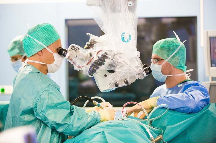 zwei Männer in grüner OP-Kleidung blicken durch ein Vergrößerungsgerät und halten medizinisches Werkzeug in den Händen